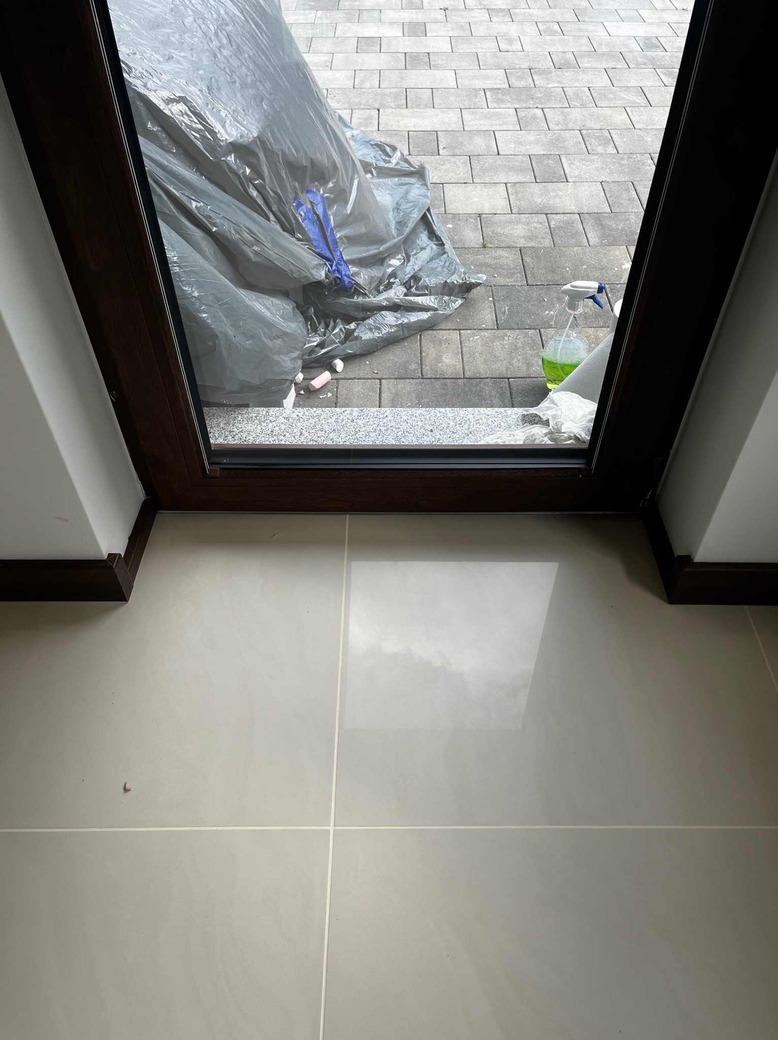 Widok przez okno balkonowe na zabezpieczony folią balkon z szarą kostką brukową. Wnętrze z jasnymi, połyskującymi płytkami. Widoczny płyn do czyszczenia.