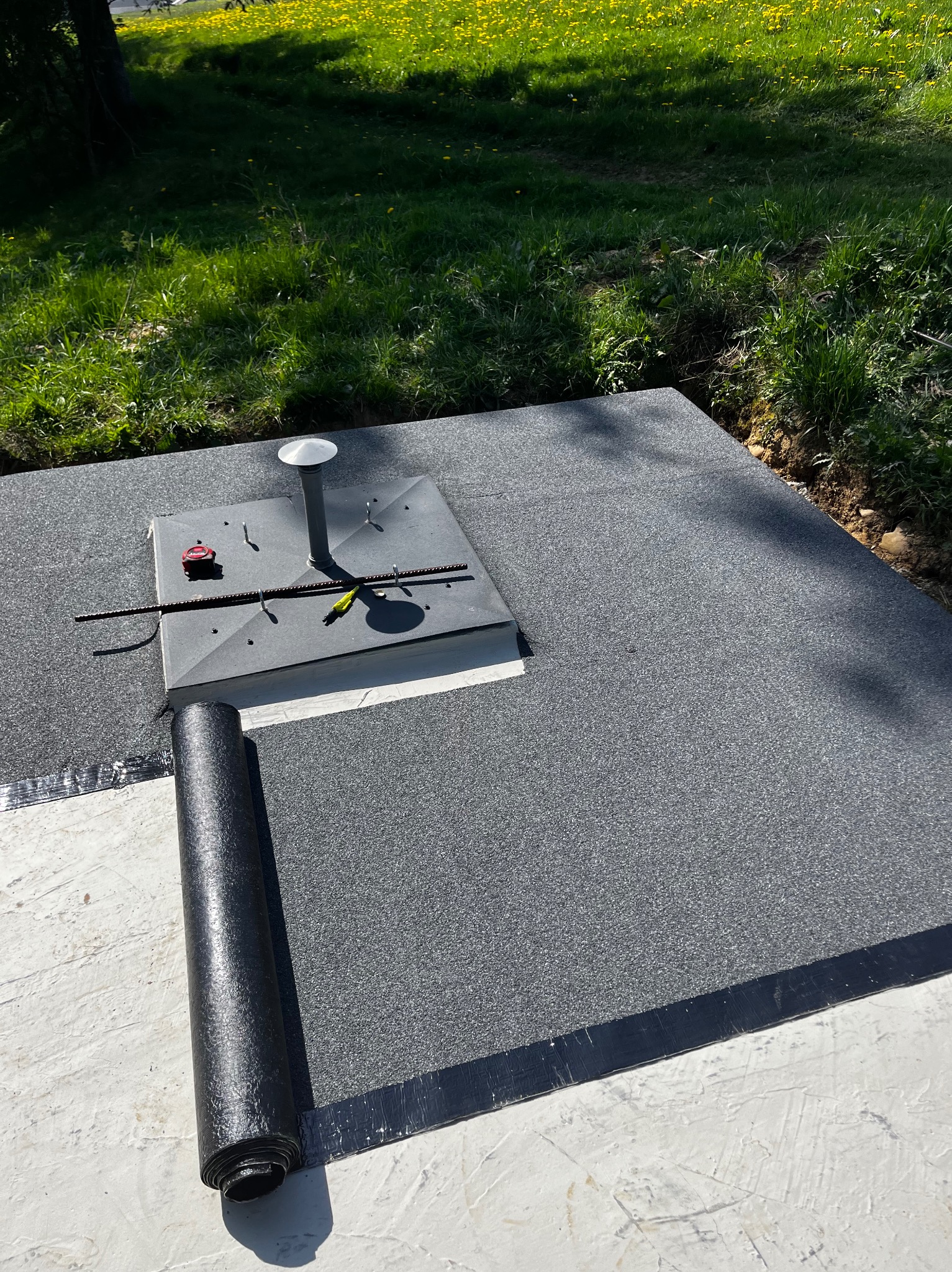 Szara papa bitumiczna na płaskim dachu, widoczny kominek wentylacyjny, narzędzia i rolka papy. Trawa i drzewa w tle.