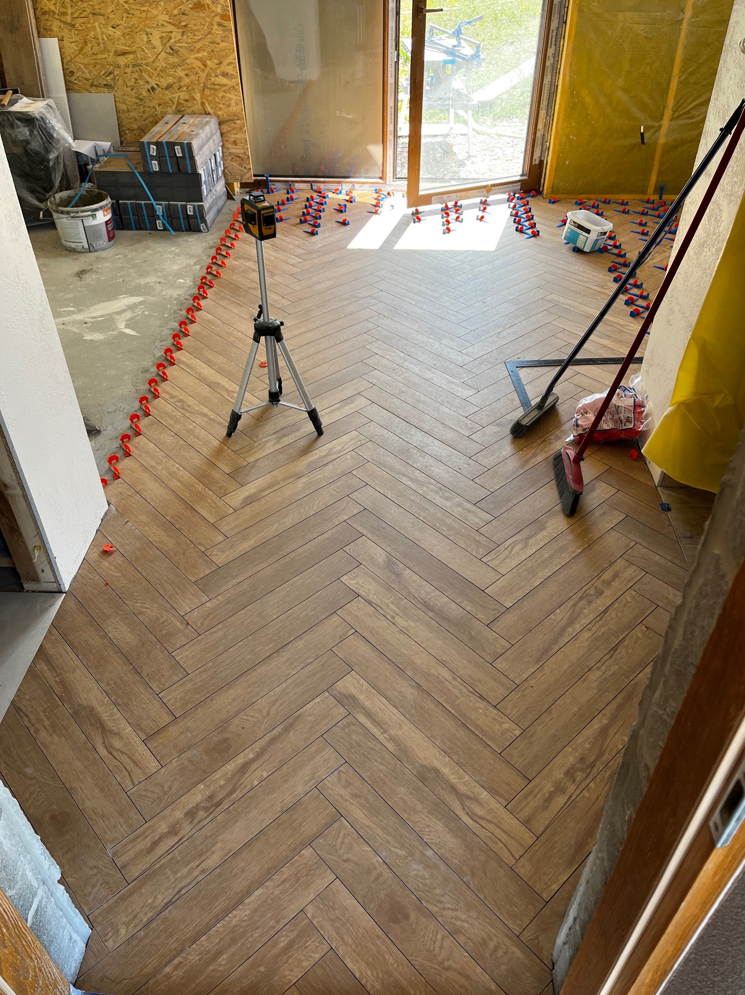 Układanie parkietu w jodełkę z użyciem poziomicy laserowej i klipsów. Widoczne płytki, narzędzia glazurnicze i surowy stan wykończenia pomieszczenia.