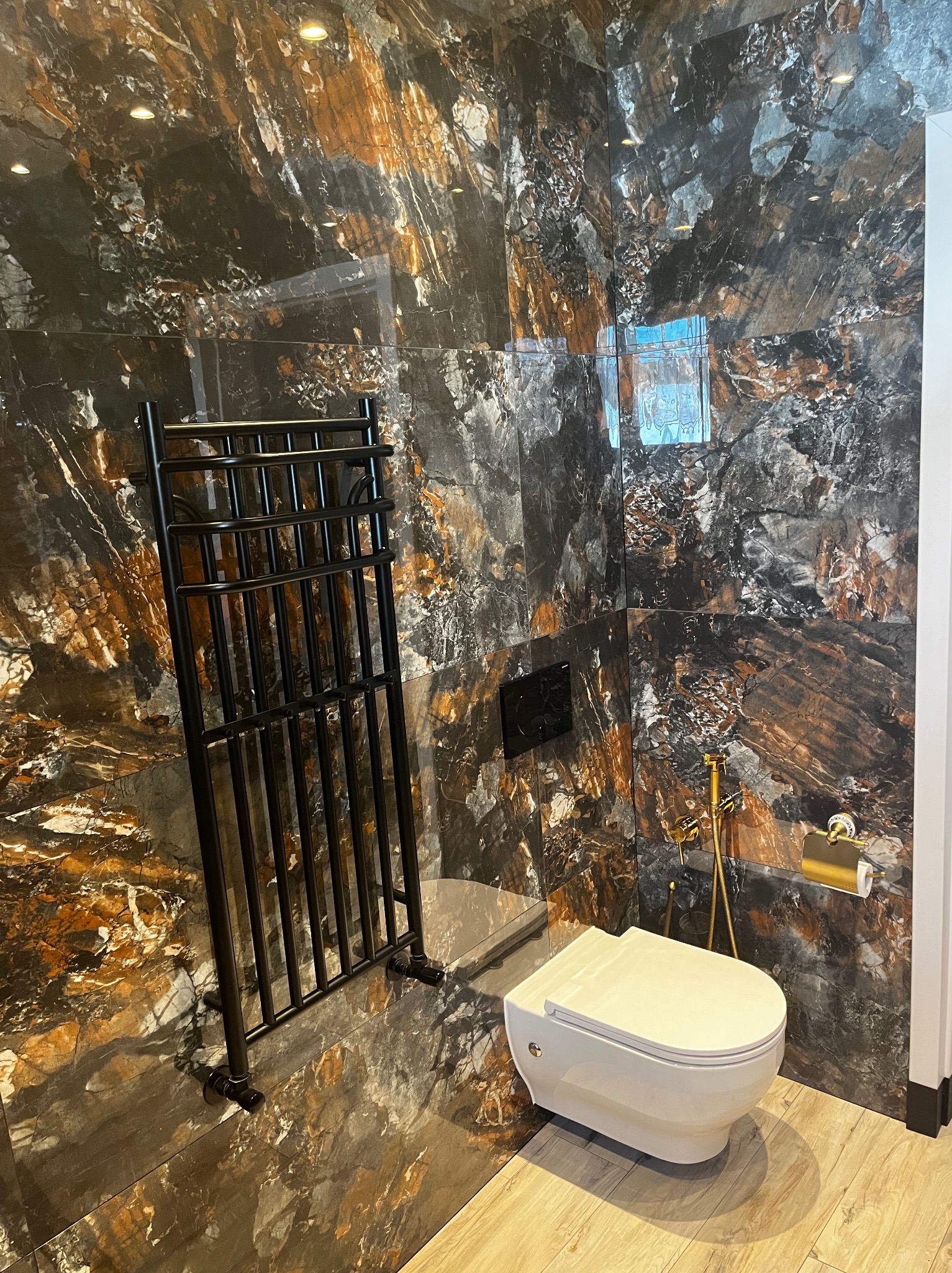 Wykończona łazienka z płytkami imitującymi marmur w odcieniach czerni i brązu. Widoczny biały sedes, czarny grzejnik drabinkowy i złote akcesoria.