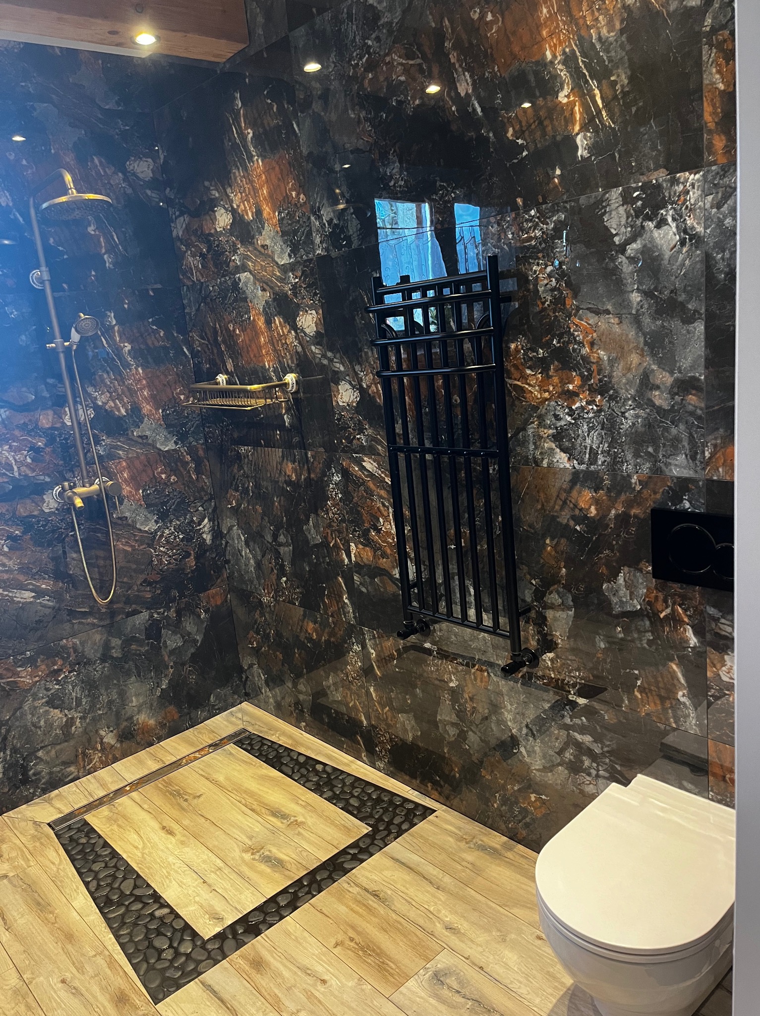 Nowoczesna łazienka z deszczownicą, grzejnikiem drabinkowym i toaletą. Ściany i podłoga wyłożone płytkami imitującymi marmur i drewno.