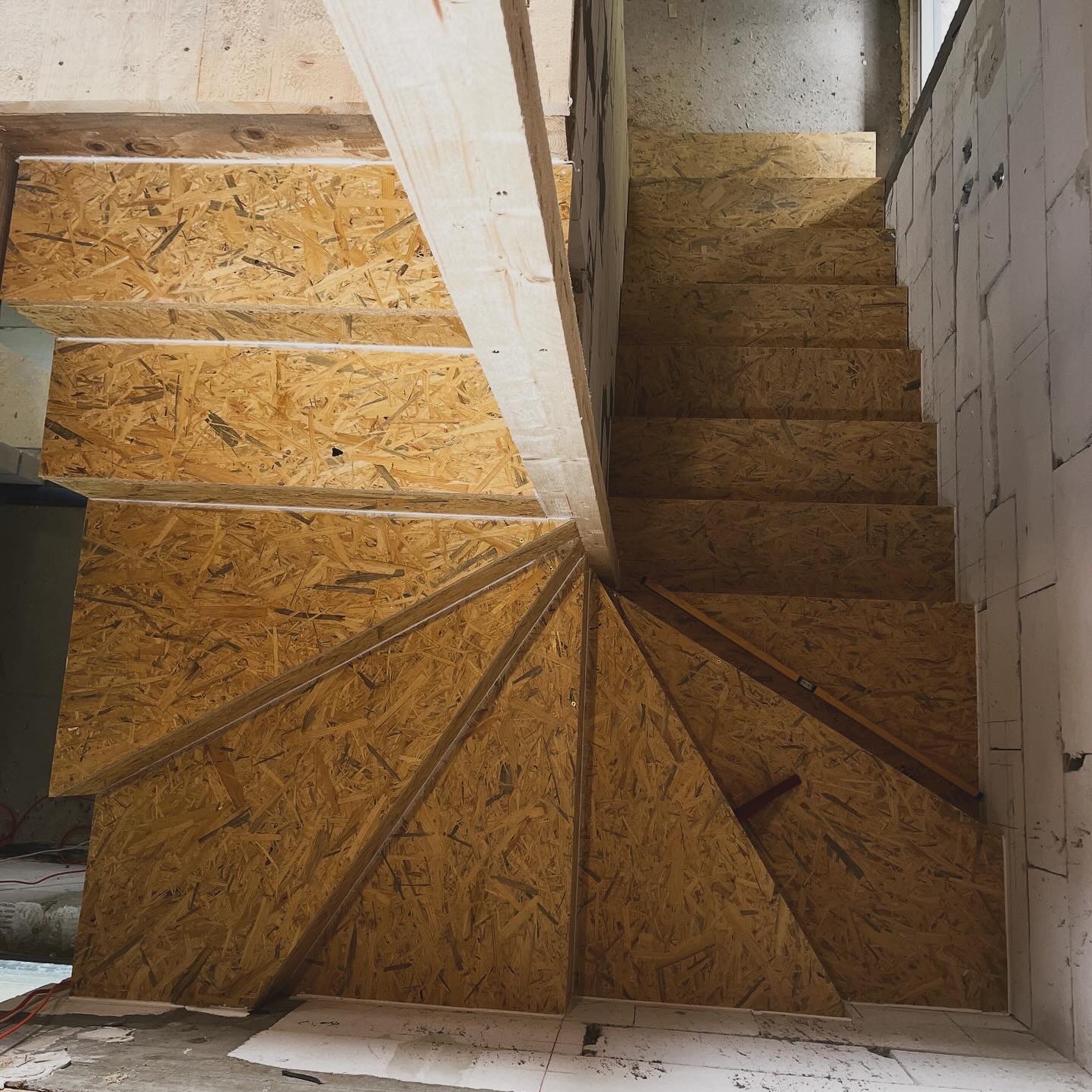 Wnętrze w trakcie budowy: drewniane schody z płyt OSB, surowy beton i bloczki. Widok z góry podkreśla spiralny kształt schodów i surowy charakter budowy.