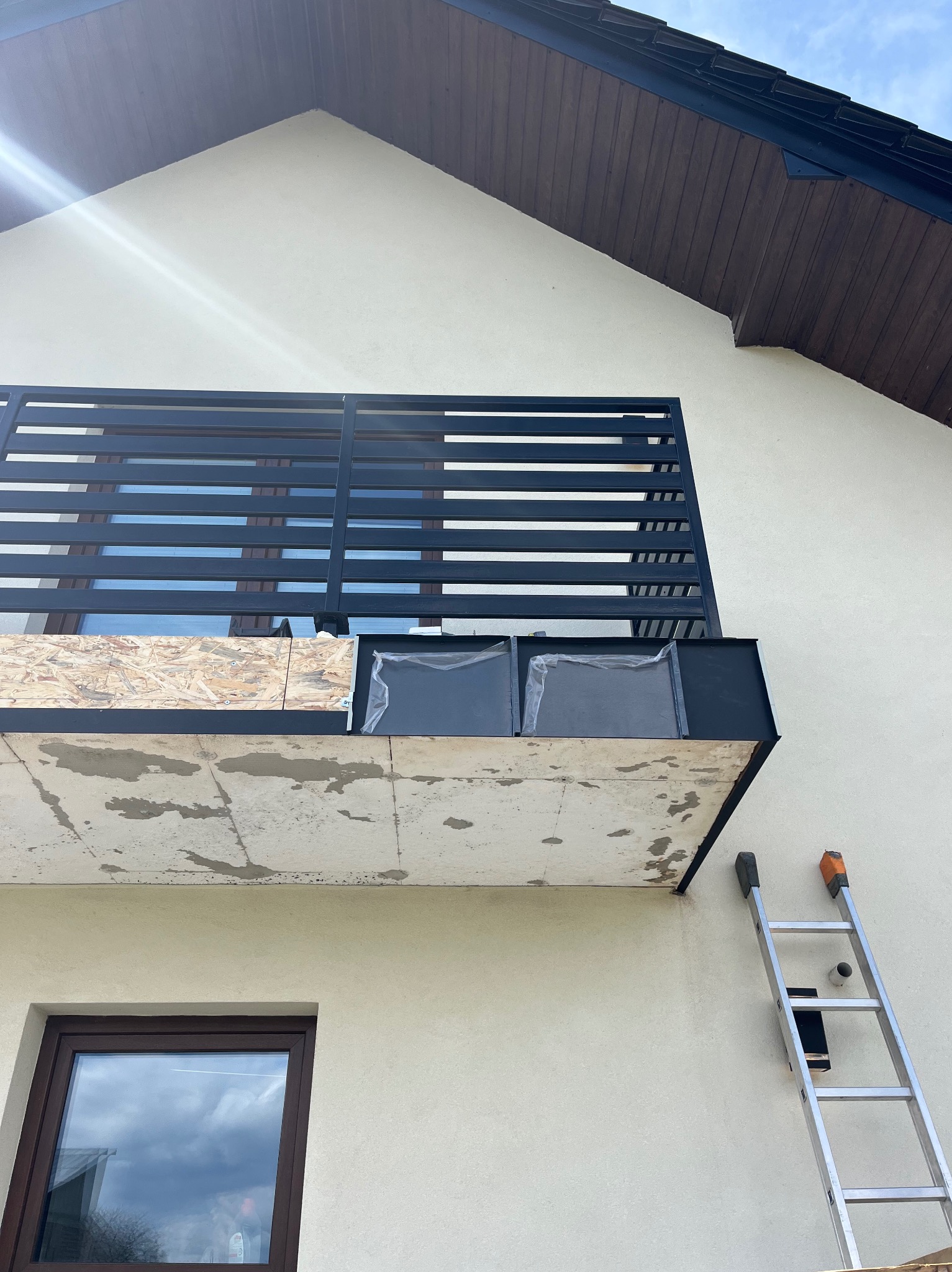 Balkon w trakcie renowacji: surowy beton, fragmenty osłony, czarna balustrada z poziomych listew. Aluminiowa drabina oparta o ścianę budynku z oknem.