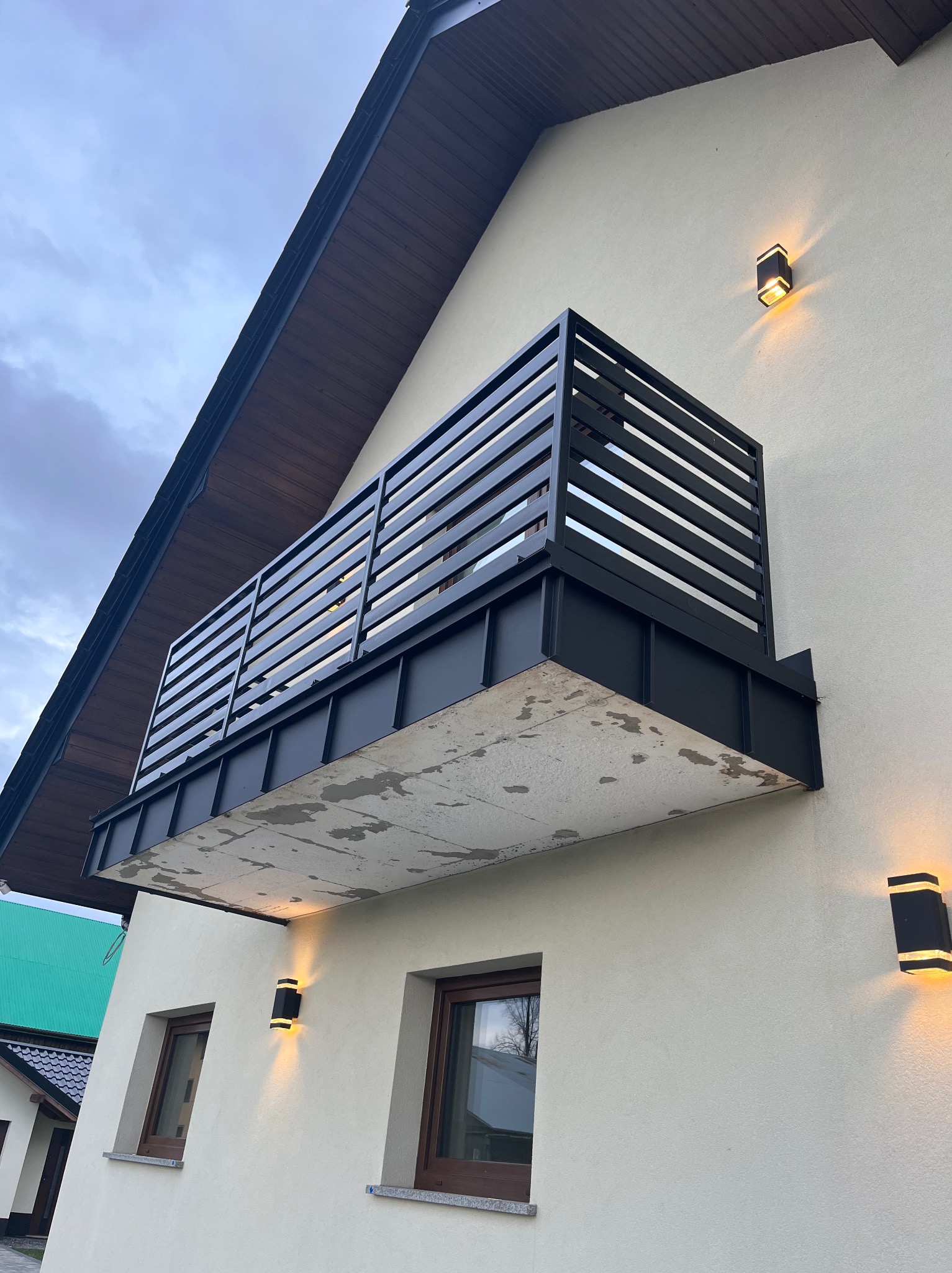 Nowoczesny balkon z czarną, poziomą balustradą na tle jasnej elewacji domu. Widoczne oświetlenie zewnętrzne i fragment dachu. Ujęcie z dołu.