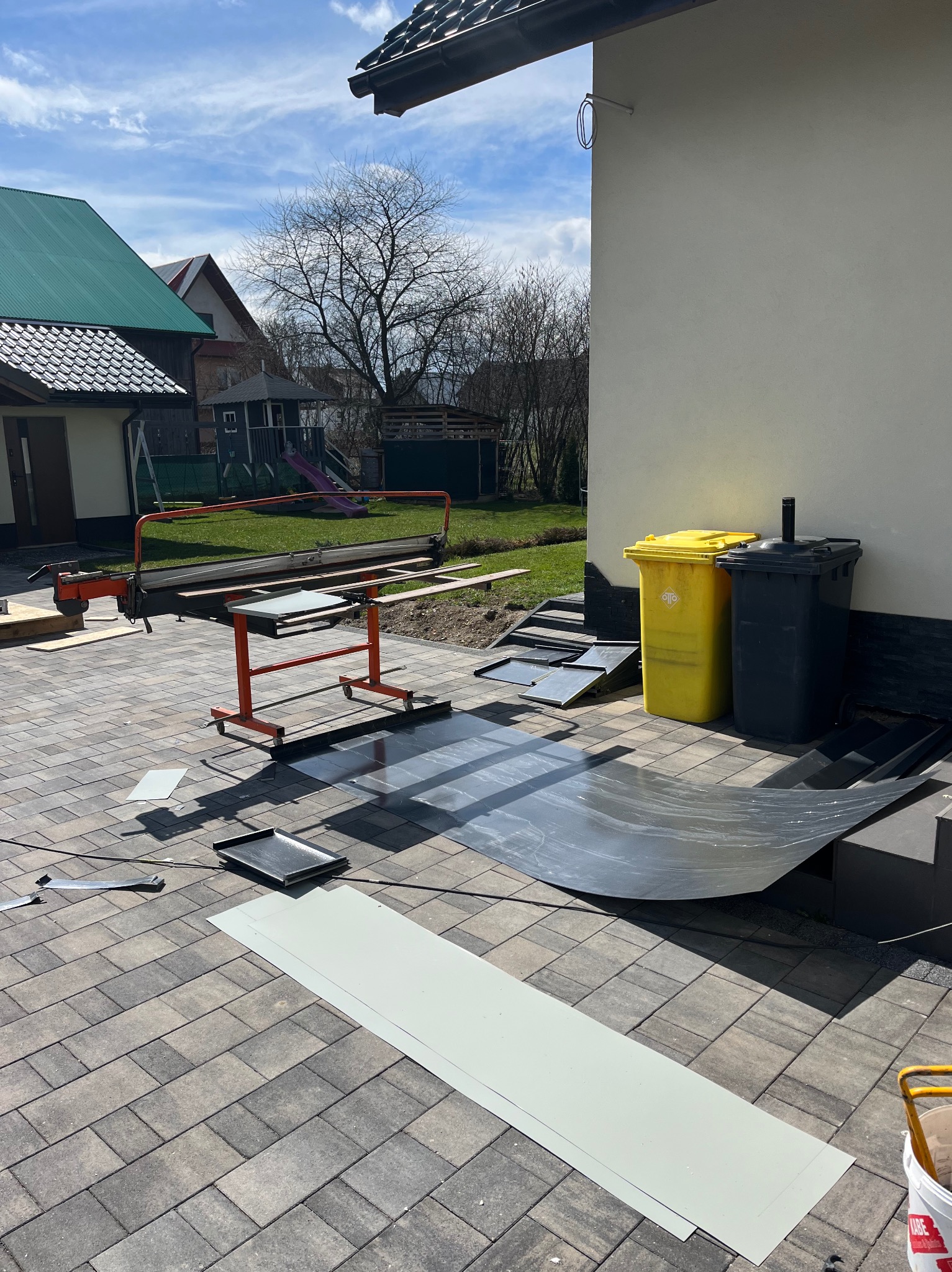 Przygotowanie blachy na zewnątrz budynku, widoczny gilotyna do cięcia blachy, arkusze blachy na kostce brukowej, żółty i czarny pojemnik na śmieci.