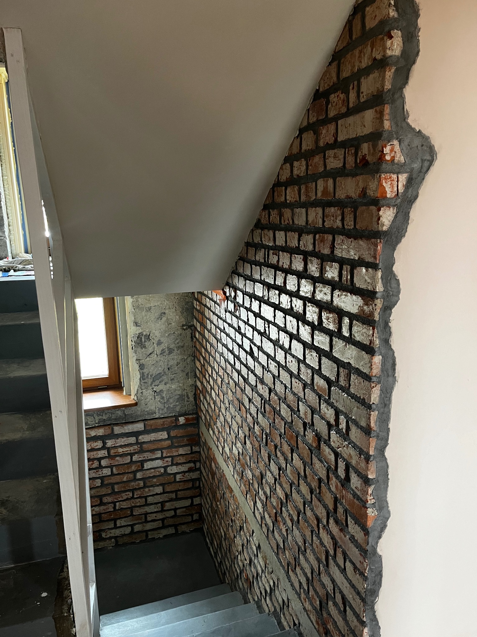 Klatka schodowa w trakcie prac wykończeniowych. Fragment ściany z cegły, surowy beton, okno. Widoczne schody i fragment metalowej konstrukcji.