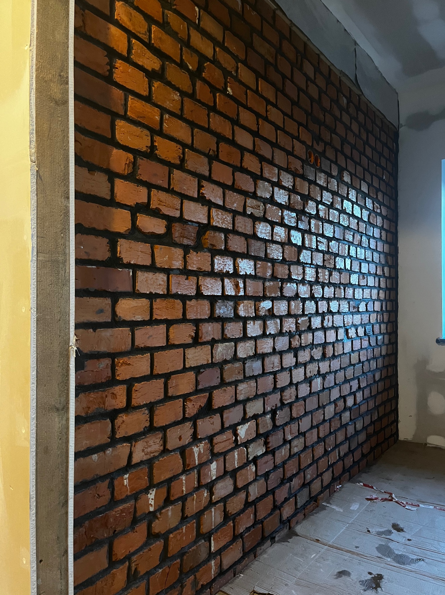 Ściana z cegły klinkierowej w surowym wnętrzu w Starym Bystrem. Widoczne niedokończone prace wykończeniowe i fragment okna. Cegła o nieregularnej fakturze.