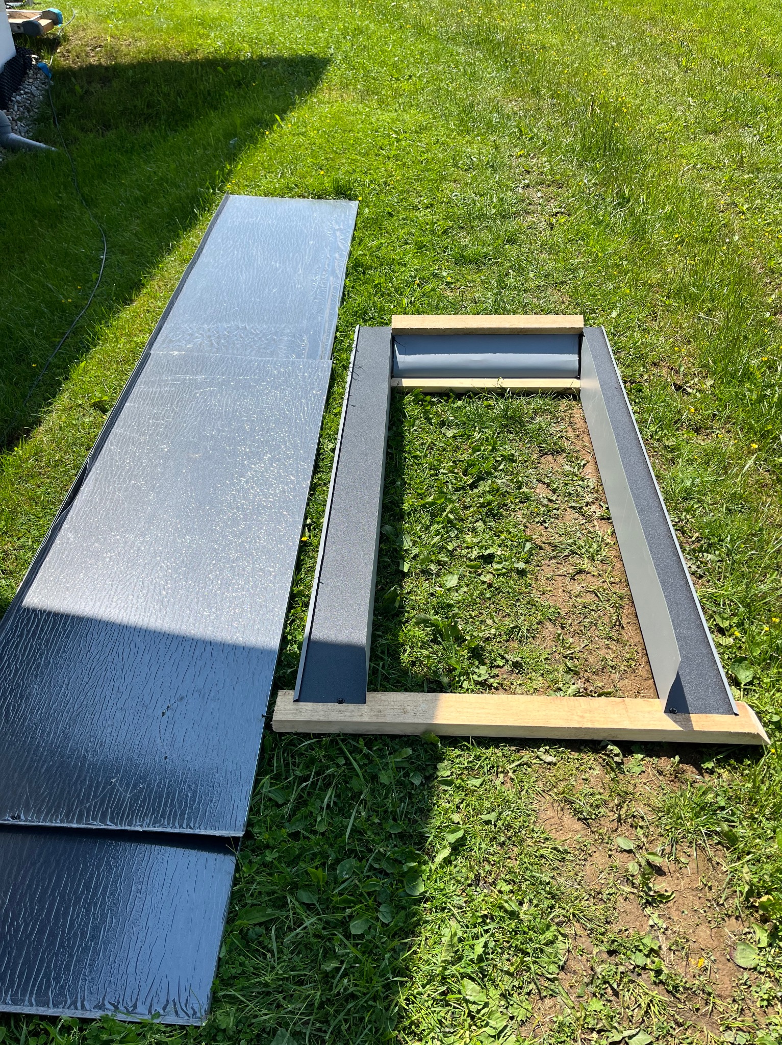 Elementy konstrukcyjne zadaszenia na trawie: blacha falista i rama z drewnianych belek, przygotowane do montażu. Widok z góry, w słoneczny dzień.