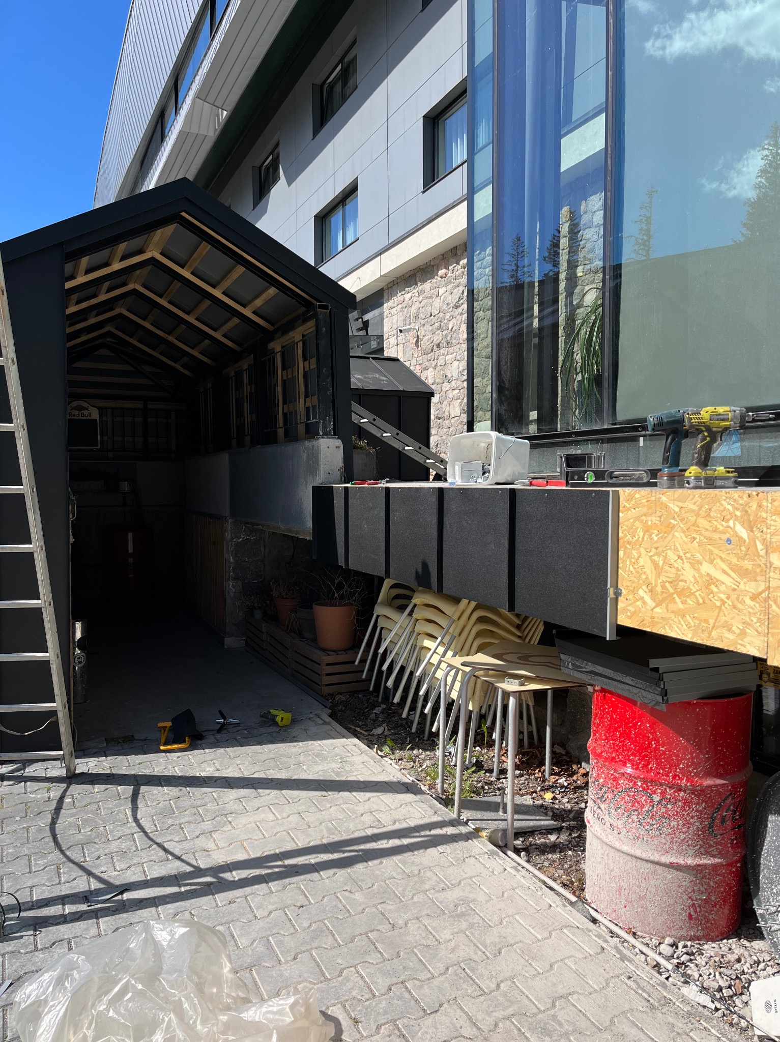 Zewnętrzna zabudowa z drewnianą konstrukcją i narzędziami. Widoczna drabina, krzesła, czerwona beczka i kostka brukowa. Prace w toku, częściowo zadaszone.