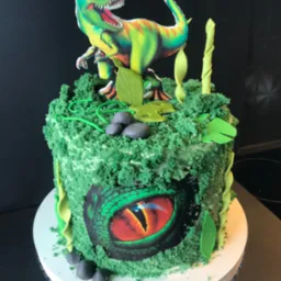 Tort urodzinowy z motywem dinozaura: zielony krem, figurka T-Rexa i oko gada. Dekoracje z liści i kamieni.
