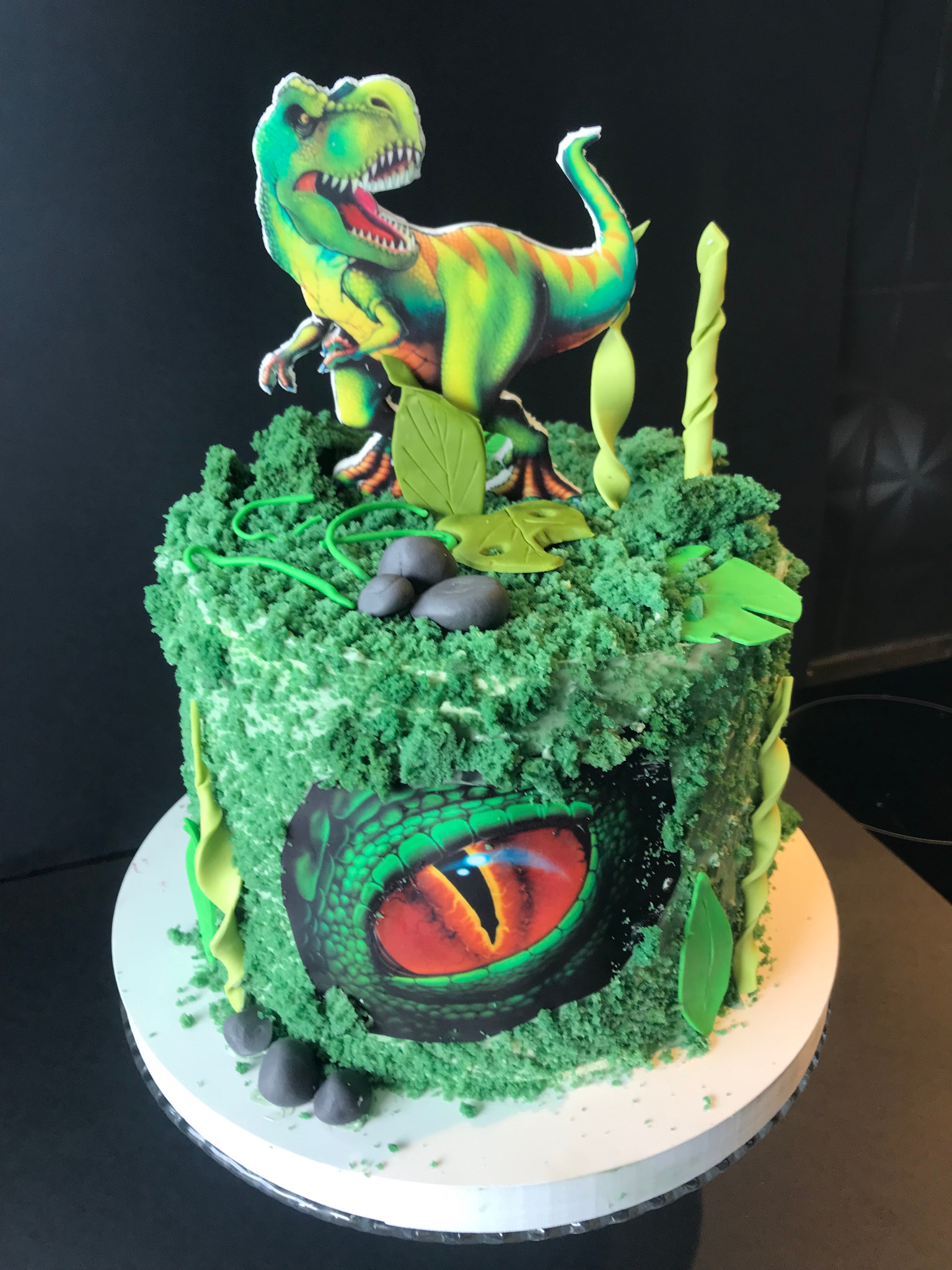 Tort urodzinowy z motywem dinozaura: zielony krem, figurka T-Rexa i oko gada. Dekoracje z liści i kamieni.