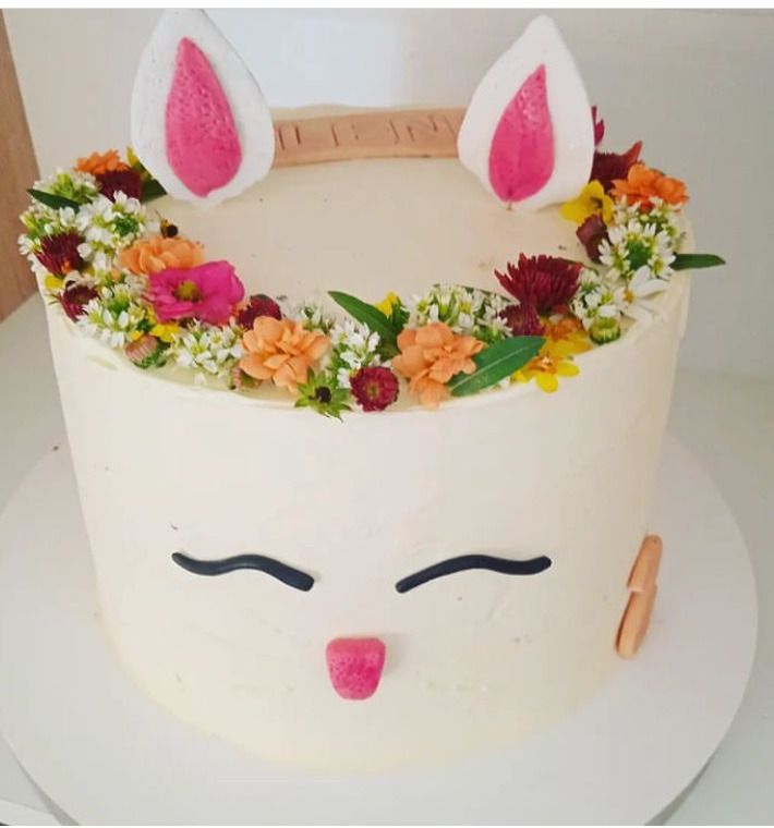 Tort w stylu kawaii z białym kremem, kwiatową dekoracją i uszami królika, umieszczony na białym talerzu. Uroczy, ręcznie robiony wypiek.