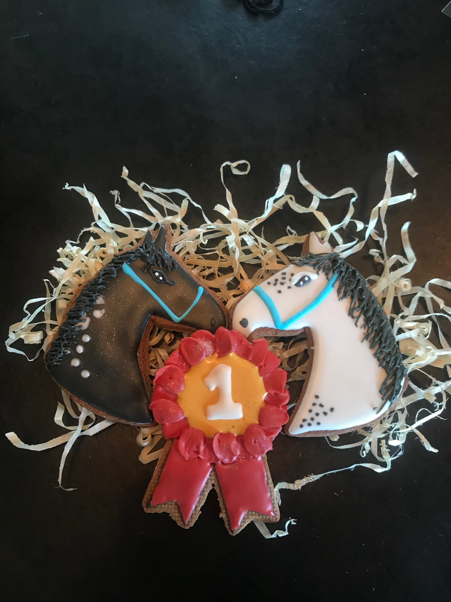 Dwa pierniki w kształcie końskich głów (biały i brązowy) oraz piernik z numerem 1, otoczone dekoracyjnym siankiem na czarnym tle. Słodki upominek.