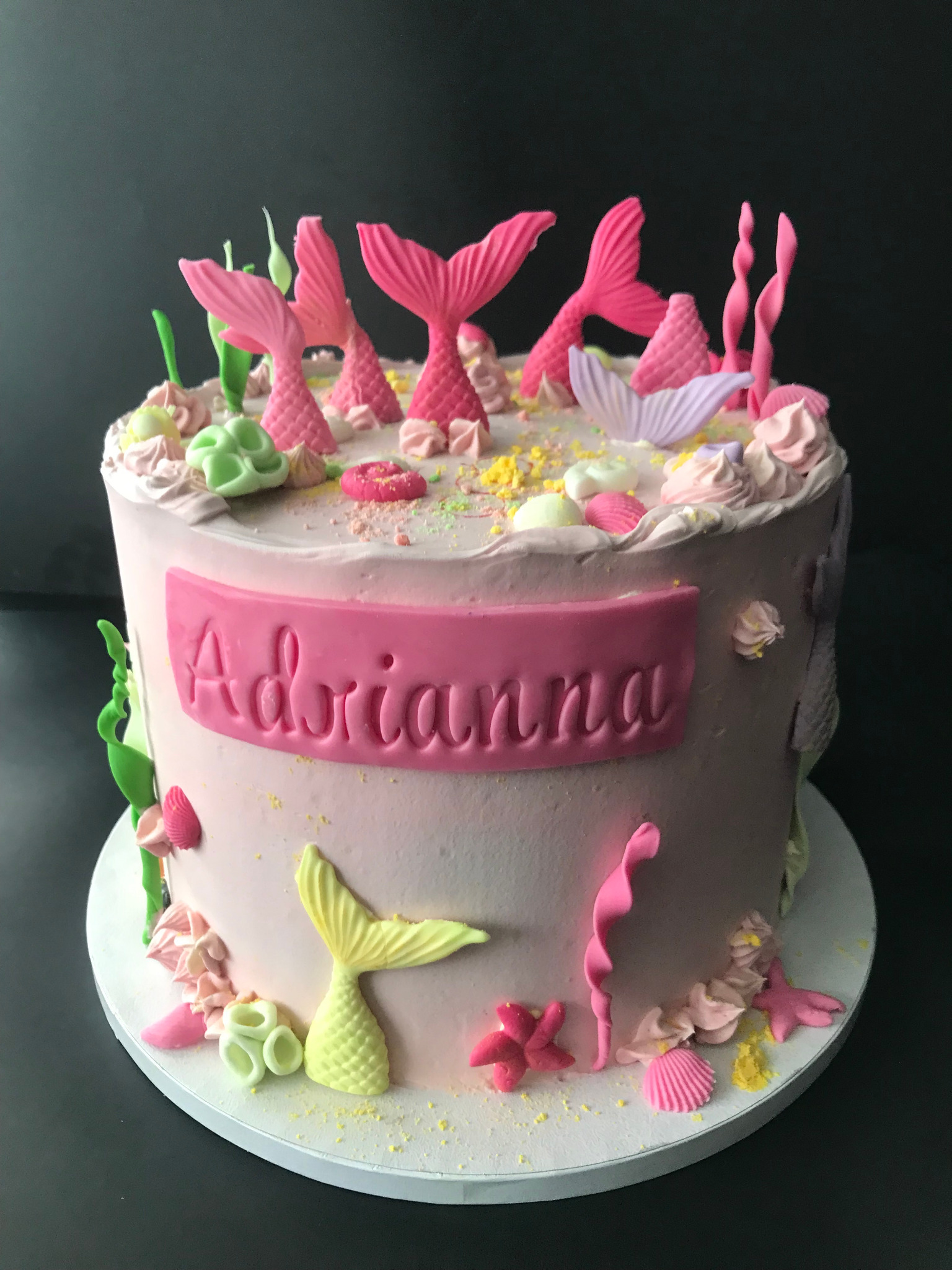 Tort urodzinowy w stylu syreny z różowym lukrem, ogonami syreny i imieniem Adrianna. Dekoracje z motywami morskimi na tle czarnego tła.