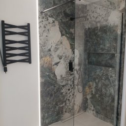 Pat-Bud - Nowoczesna kabina prysznicowa z czarnymi detalami i marmurową ścianą, oświetlona listwą LED. Grzejnik drabinkowy w kolorze czarnym na białej ścianie.