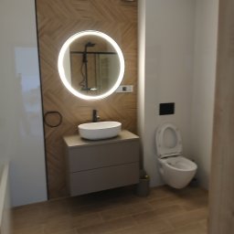Pat-Bud - Nowoczesna łazienka z okrągłym lustrem LED, drewnianymi panelami i beżowymi płytkami podłogowymi. Wisząca toaleta i minimalistyczna umywalka na szafce.
