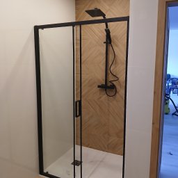 Pat-Bud - Nowoczesna kabina prysznicowa z czarnymi profilami i deszczownicą na tle ściany wyłożonej płytkami imitującymi drewno. Widok na wykończoną łazienkę.