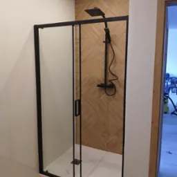 Nowoczesna kabina prysznicowa z czarnymi profilami i deszczownicą na tle ściany wyłożonej płytkami imitującymi drewno. Widok na wykończoną łazienkę.