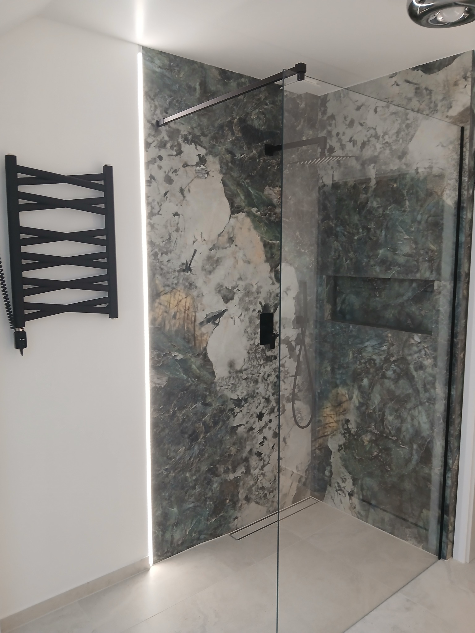 Nowoczesna kabina prysznicowa z czarnymi detalami i marmurową ścianą, oświetlona listwą LED. Grzejnik drabinkowy w kolorze czarnym na białej ścianie.