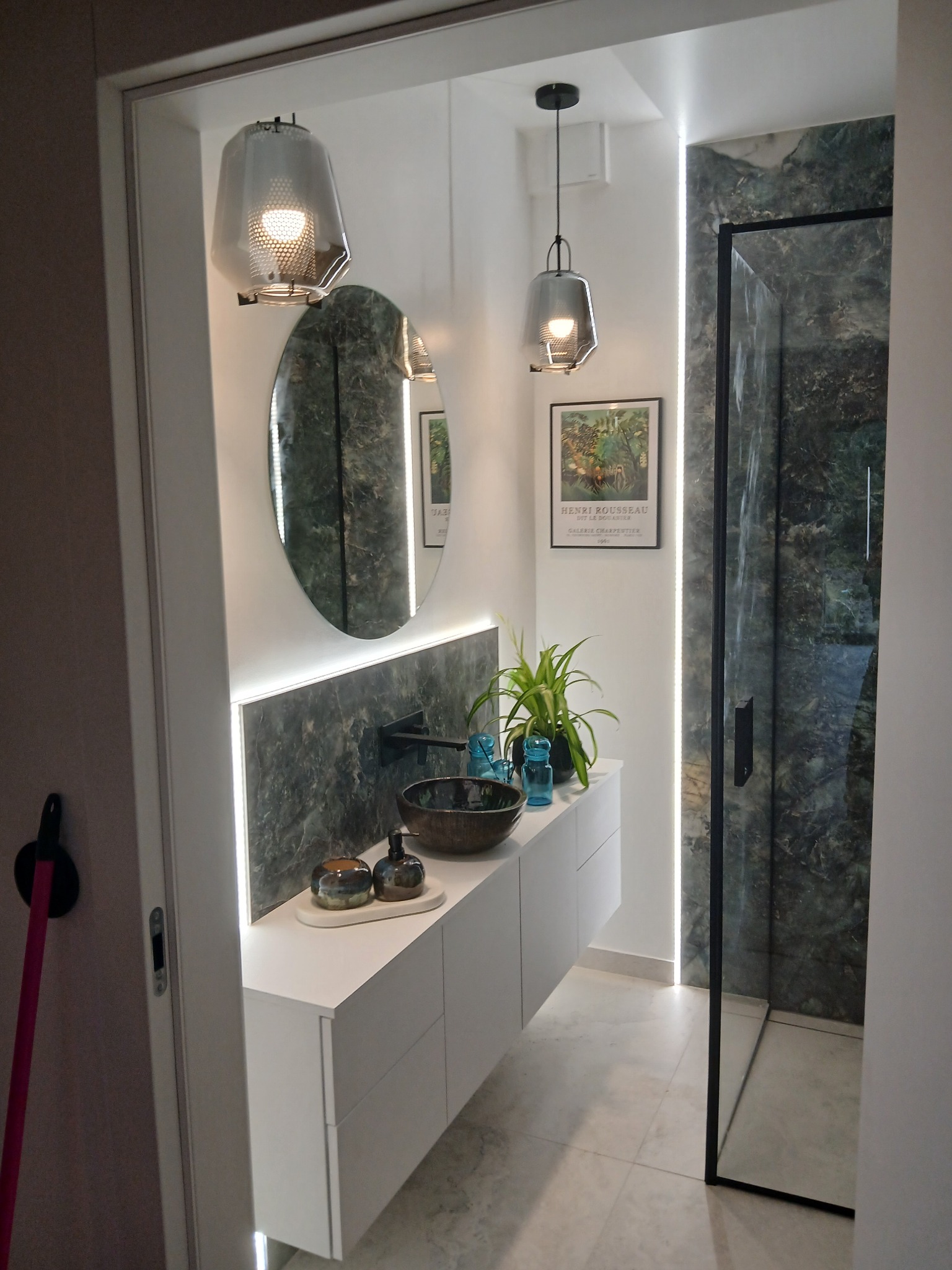 Nowoczesna łazienka z owalnym lustrem, marmurową ścianą i szklaną kabiną prysznicową. Minimalistyczne oświetlenie i rośliny dodają wnętrzu elegancji.