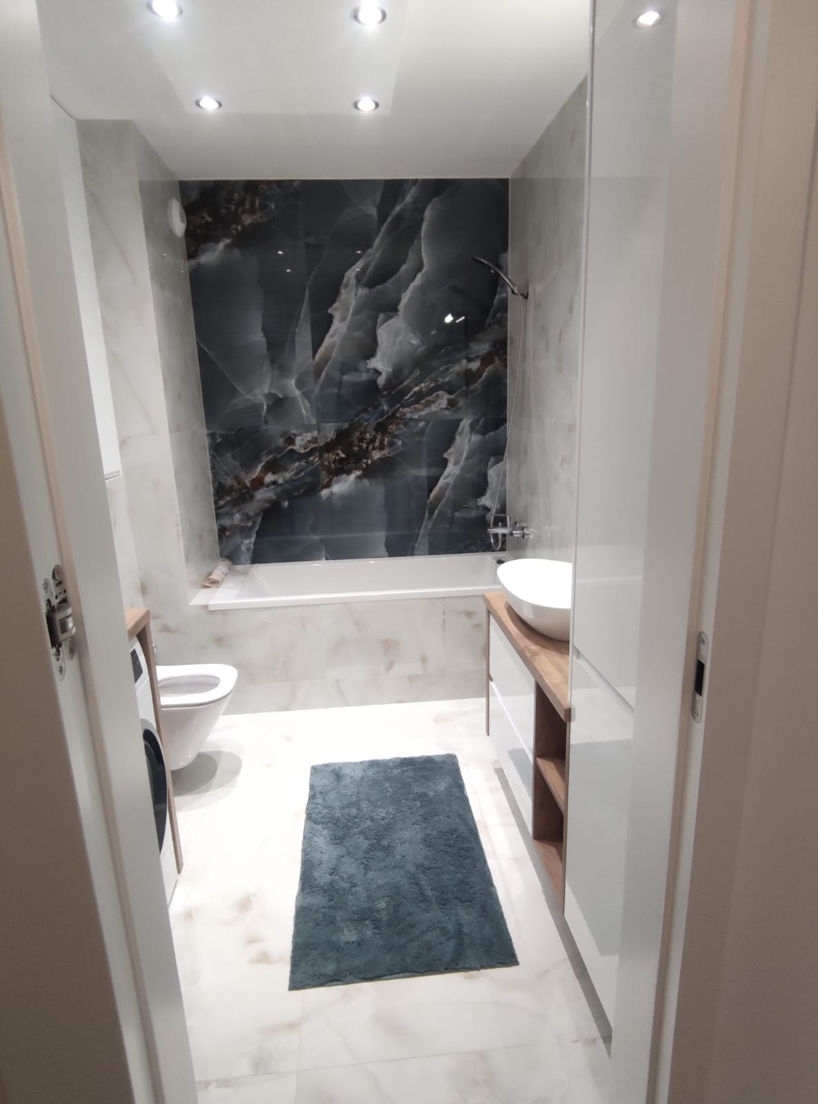 Wykończona łazienka z marmurową ścianą, białą armaturą i drewnianymi akcentami. Widok zza uchylonych drzwi, dywanik na podłodze.
