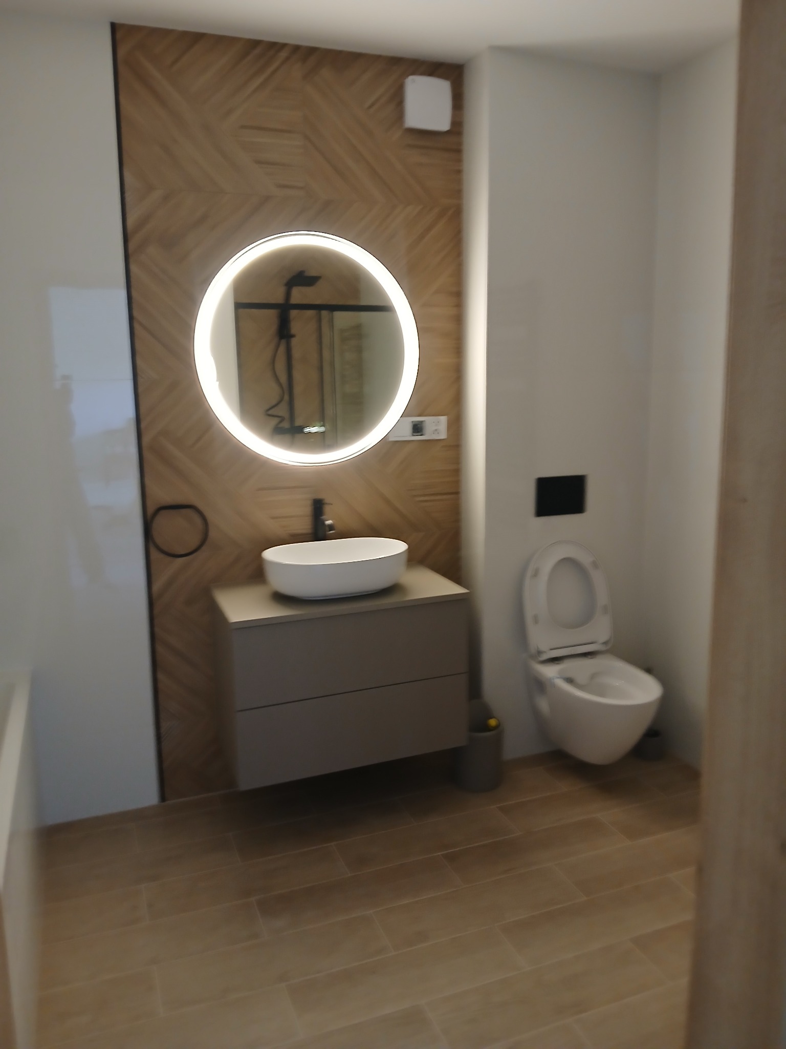 Nowoczesna łazienka z okrągłym lustrem LED, drewnianymi panelami i beżowymi płytkami podłogowymi. Wisząca toaleta i minimalistyczna umywalka na szafce.