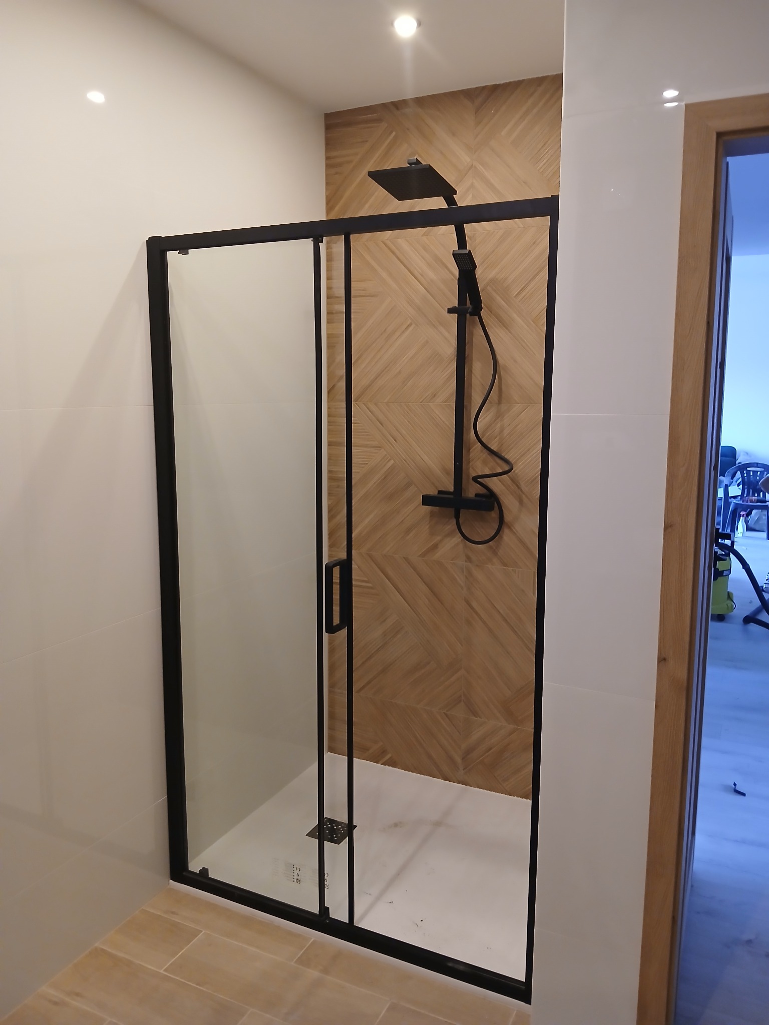 Nowoczesna kabina prysznicowa z czarnymi profilami i deszczownicą na tle ściany wyłożonej płytkami imitującymi drewno. Widok na wykończoną łazienkę.