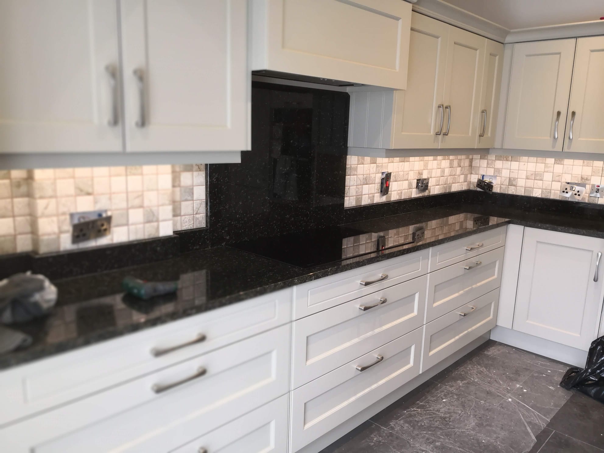 Kuchnia z białymi szafkami, czarnym blatem i mozaiką nad blatem. Widoczne gniazdka elektryczne i narzędzie na blacie. Podłoga w szarym kolorze.