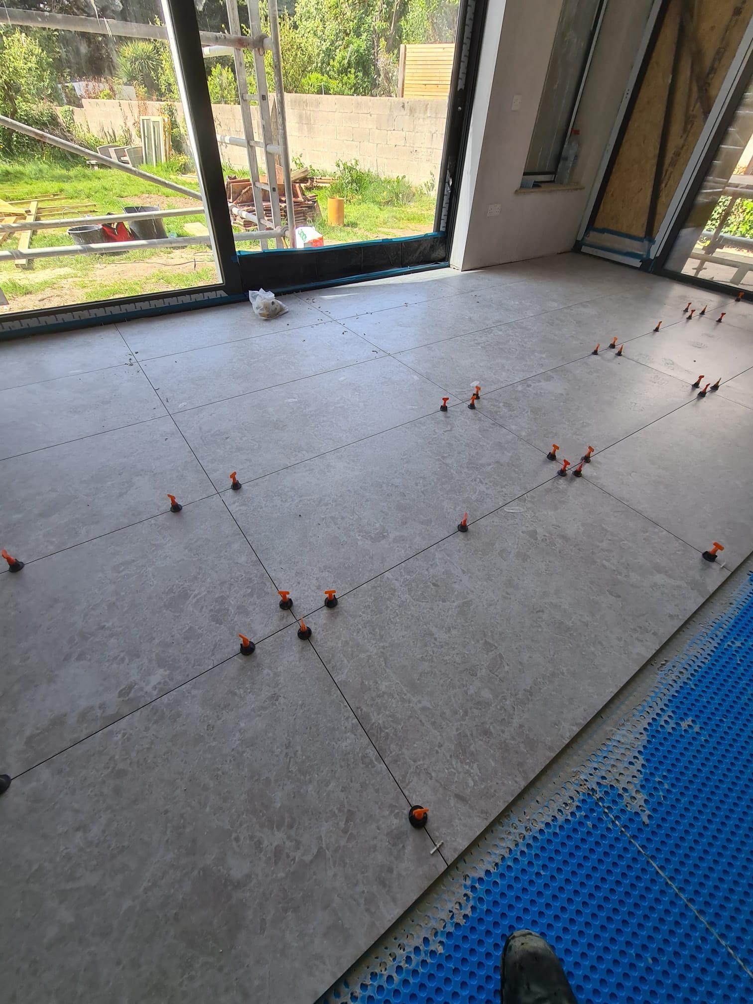 Układanie szarych płytek podłogowych z użyciem poziomujących klipsów w jasnym wnętrzu z dużymi oknami i widokiem na zewnątrz. Fragment niebieskiej maty pod płytki.