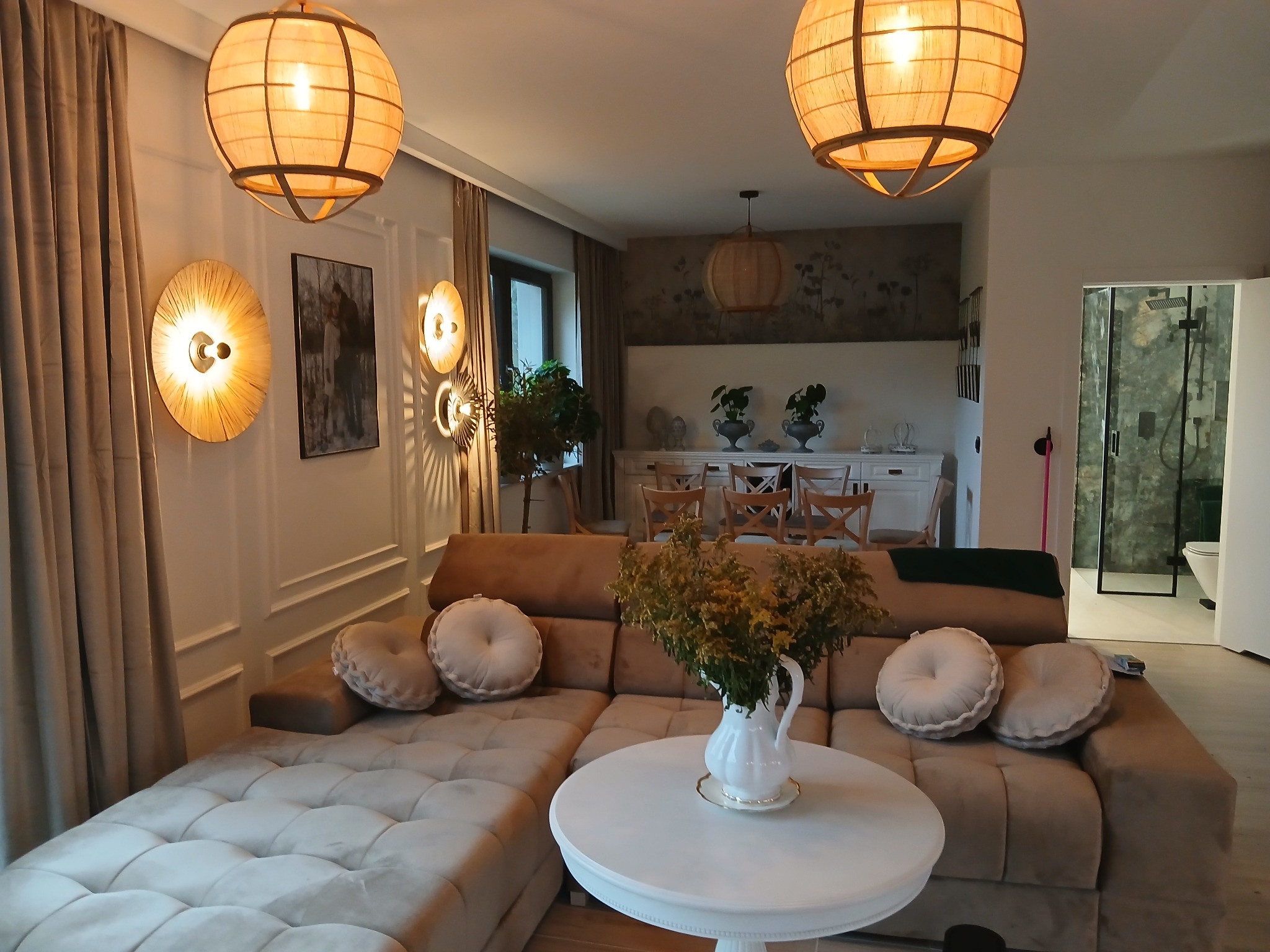 Stylowe wnętrze salonu z narożnikiem, okrągłym stolikiem i dekoracyjnym oświetleniem. W tle widoczny fragment łazienki z kabiną prysznicową.