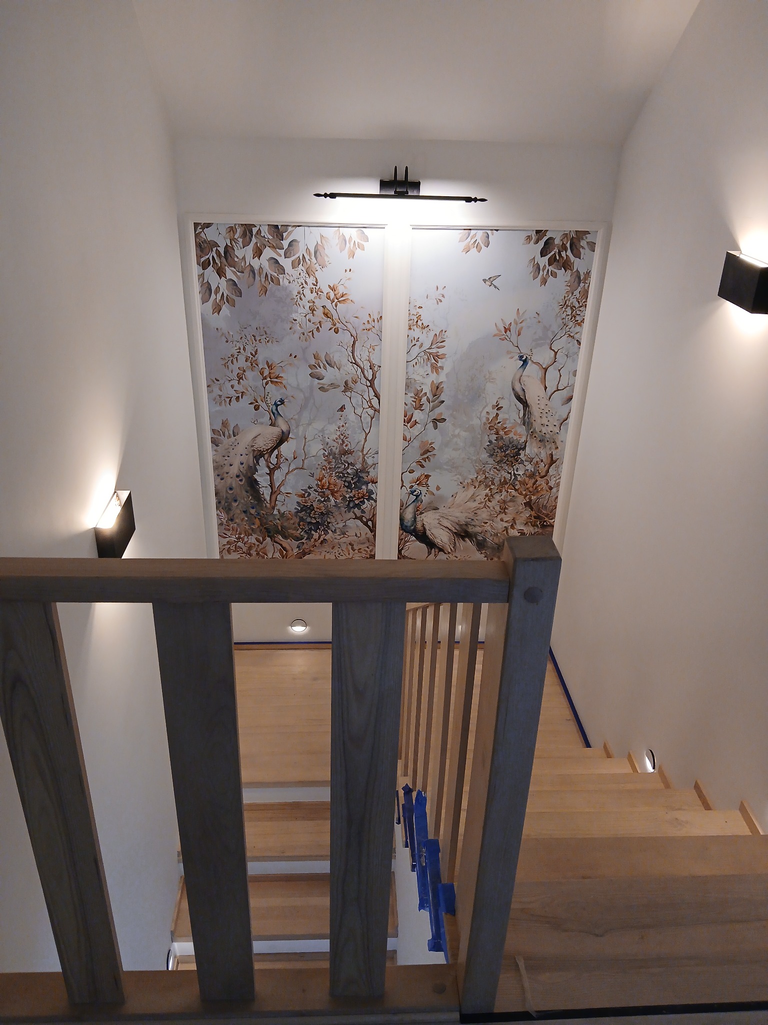 Klatka schodowa z drewnianymi schodami i balustradą. Na ścianie fototapeta z motywem pawia, oświetlona kinkietami. Widok z góry, na drewniane schody.
