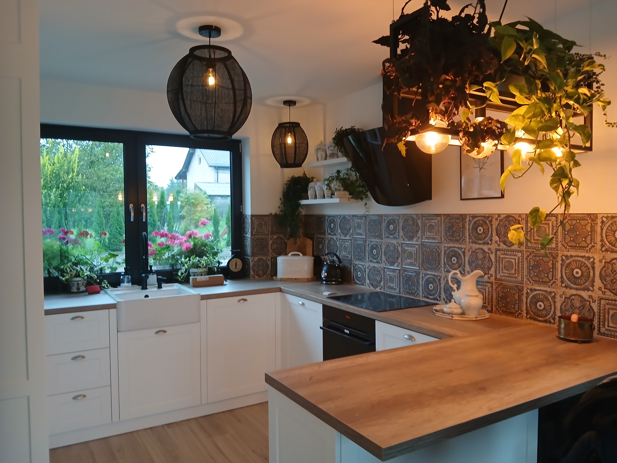 Jasna kuchnia w stylu eklektycznym: białe szafki, drewniany blat i ozdobne płytki. Okno z kwiatami i oryginalne lampy tworzą przytulny klimat wnętrza.