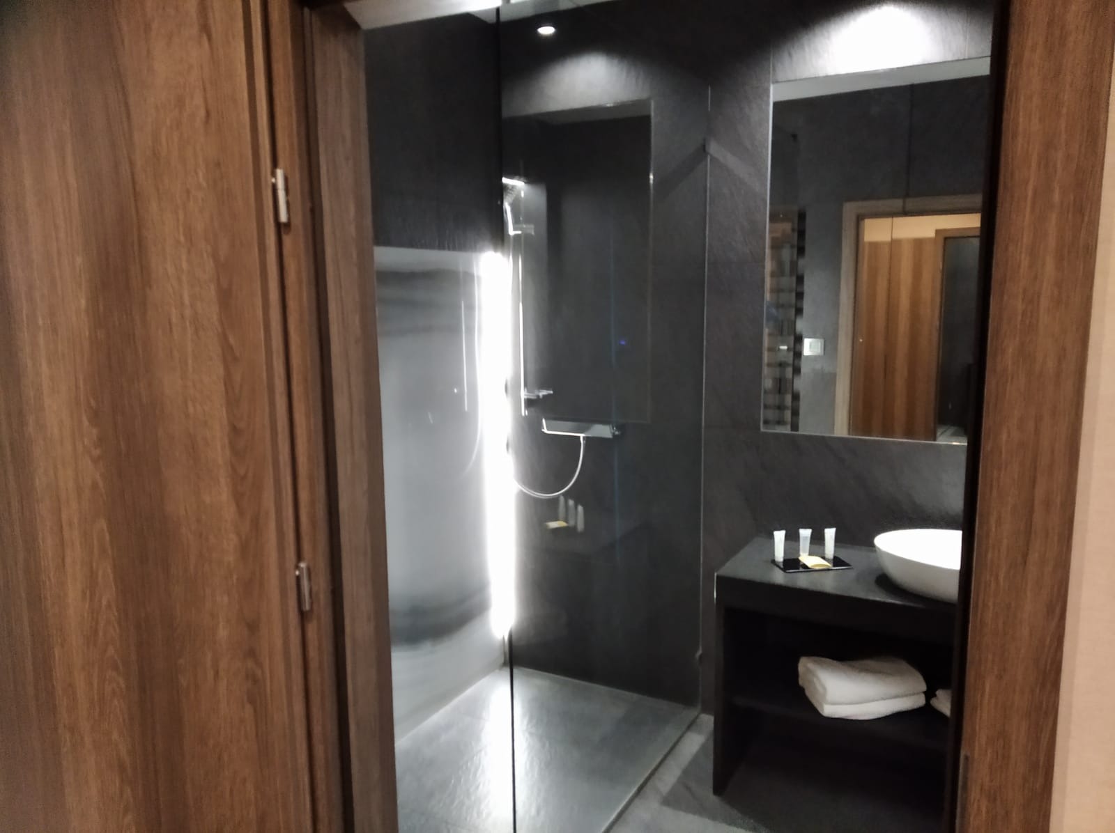 Nowoczesna łazienka z kabiną prysznicową typu walk-in, umywalką nablatową i dużym lustrem w minimalistycznym stylu. Ciemna kolorystyka podkreśla elegancję wnętrza.