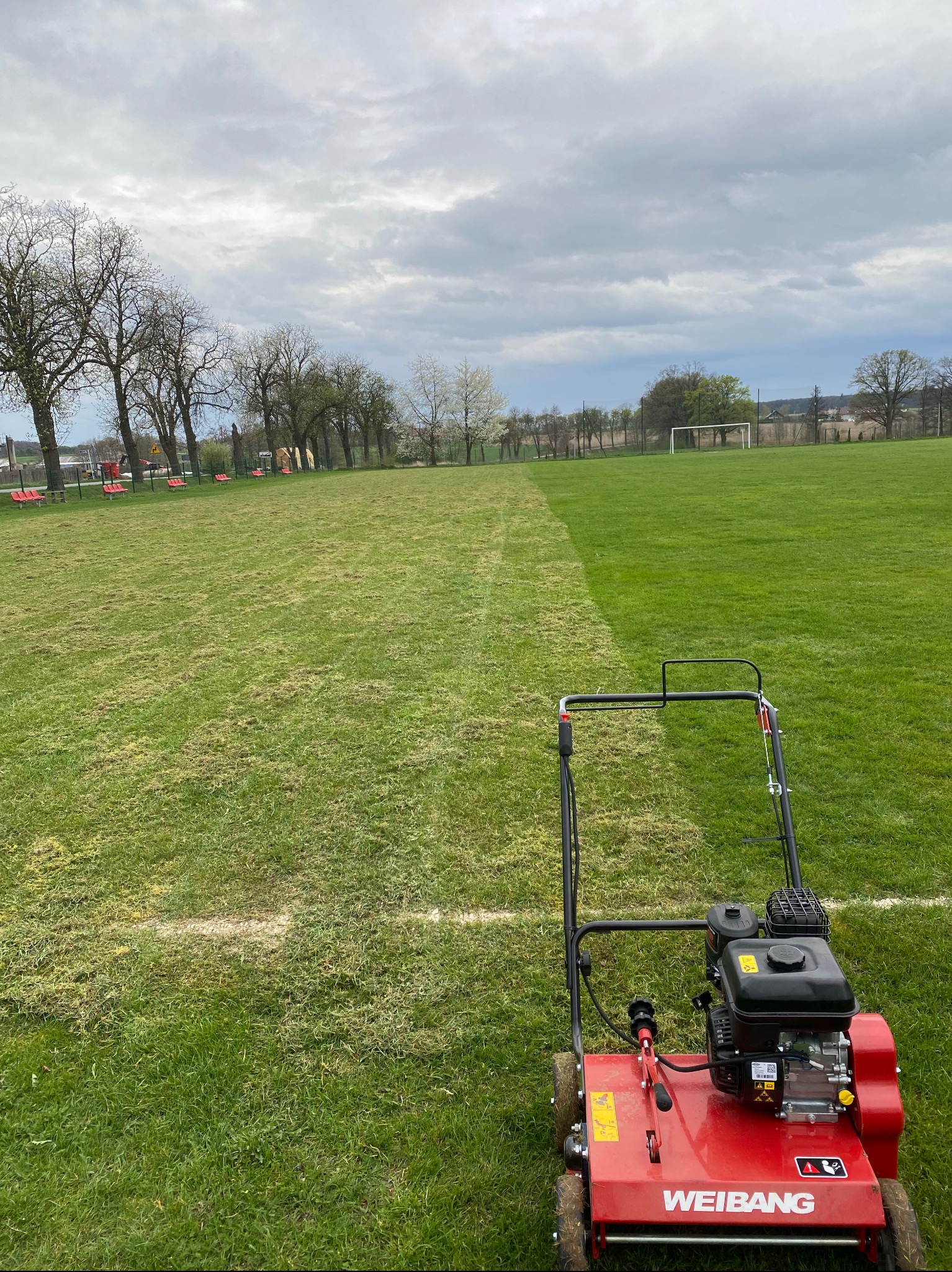 Czerwona kosiarka WEIBANG na boisku, połowa trawy skoszona, widoczna linia podziału. W tle drzewa i bramka. Pochmurne niebo.