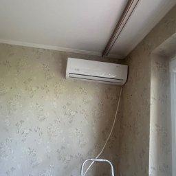 Klima-art s.c Bartosz Kopka Lilianna Szyszka-Kazimierska - Biały klimatyzator na ścianie w pokoju z kwiecistą tapetą, widoczny fragment drabiny i jednostka zewnętrzna za oknem. Standardowy montaż klimatyzacji.