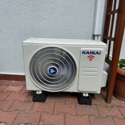 Klima-art s.c Bartosz Kopka Lilianna Szyszka-Kazimierska - Zewnętrzna jednostka klimatyzacji Kaisai na czerwonej kostce brukowej, wsparta na czarnych podkładkach antywibracyjnych, przy ścianie z drewnianą konstrukcją i donicą.