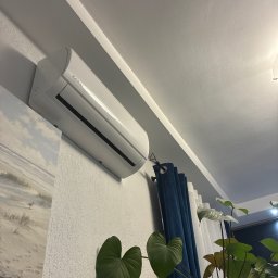 Klima-art s.c Bartosz Kopka Lilianna Szyszka-Kazimierska - Biała klimatyzacja Kaisai na ścianie nad obrazem plaży, z niebieskimi zasłonami i zielonymi roślinami w tle, tworząca domową atmosferę.