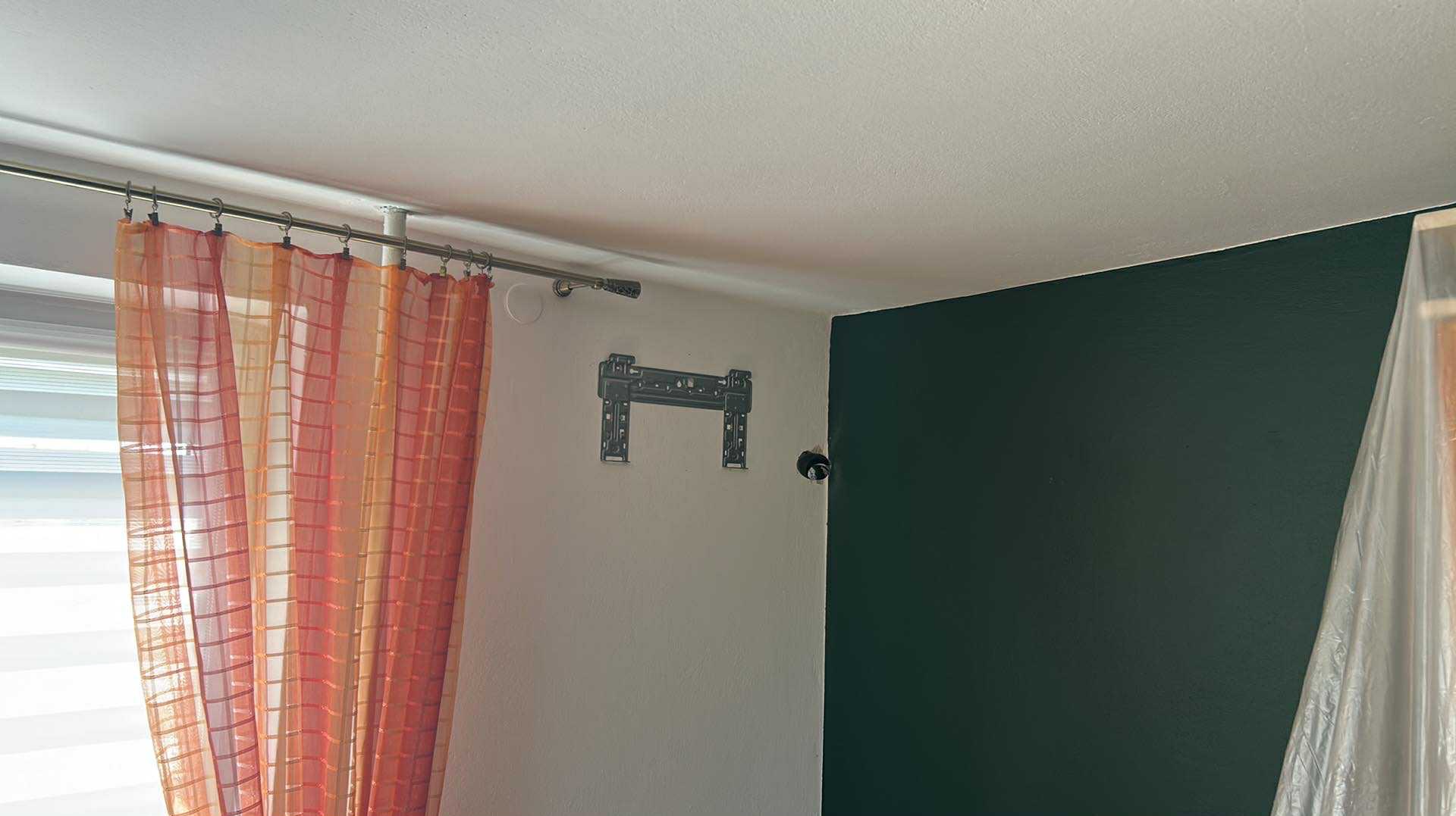 Ściana z zamontowanym uchwytem TV, zasłonięta folią część ściany pomalowana na zielono, okno z pomarańczową zasłoną w tle. Przygotowanie do montażu klimatyzacji.