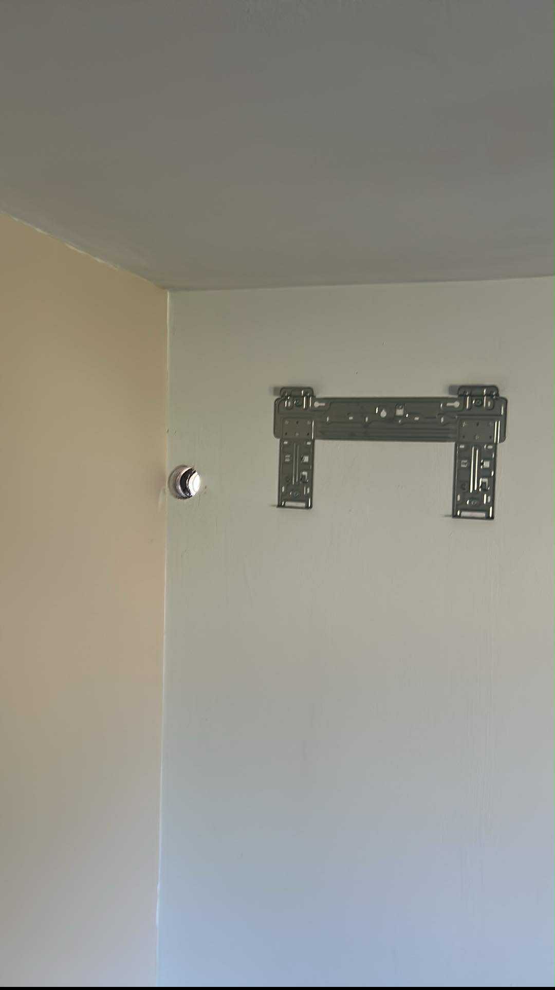 Metalowy uchwyt klimatyzatora zamontowany na ścianie, obok otworu na przewody. Widok na ścianę i sufit w jasnych odcieniach. Przygotowanie do instalacji klimatyzacji.