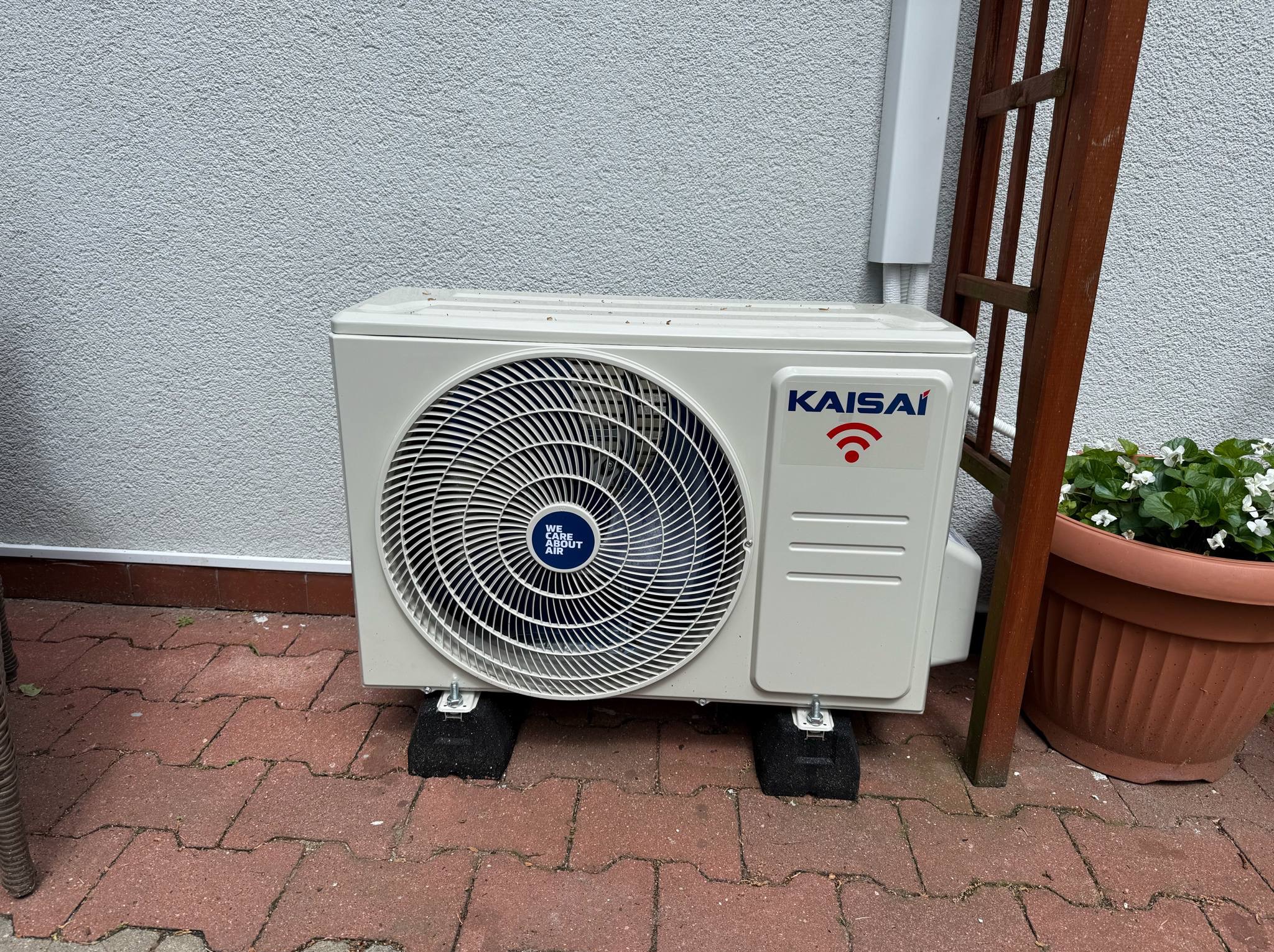 Zewnętrzna jednostka klimatyzacji Kaisai na czerwonej kostce brukowej, wsparta na czarnych podkładkach antywibracyjnych, przy ścianie z drewnianą konstrukcją i donicą.