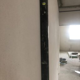 PROremont - Pionowy poziom na ścianie w surowym wnętrzu. Widoczne ślady po gładzi i szpachlowaniu. W tle okno i podłoga z materiałami budowlanymi.