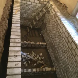 Wnętrze w trakcie prac murarskich, ściany z białej cegły, drabina oparta o ścianę, gruz i narzędzia na podłodze. Widok z góry, surowe wykończenie.