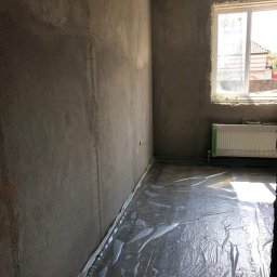 PROremont - Pokój w surowym stanie: szare ściany po tynkowaniu, okno z grzejnikiem, podłoga zabezpieczona folią. Przygotowanie do dalszych prac wykończeniowych.