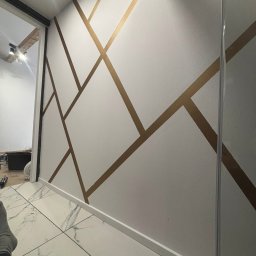 PROremont - Biała ściana z geometrycznym wzorem w złotym kolorze. Podłoga wyłożona płytkami imitującymi marmur. Widoczne detale wykończenia wnętrza.