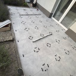 PROremont - Układanie szarych płytek na zewnątrz budynku z użyciem poziomicy i krzyżyków dystansowych. Widoczne narzędzia i fragment buta glazurnika.