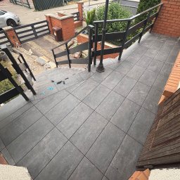 PROremont - Ułożone grafitowe płytki na schodach zewnętrznych z czarną balustradą. Widoczne narzędzia glazurnicze i fragment butów osoby fotografującej. Tło stanowi zabudowa miejska.