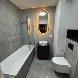 PROremont - Nowoczesna łazienka w szarościach: wanna z parawanem, podwieszana toaleta i umywalka z podświetlanym lustrem. Marmurowe płytki na ścianach i podłodze.