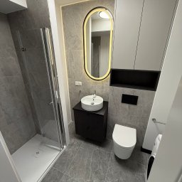PROremont - Nowoczesna łazienka w szarościach: kabina prysznicowa, umywalka na szafce, toaleta, podświetlane lustro owalne, płytki imitujące marmur, minimalistyczne szafki.