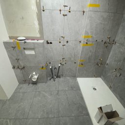 PROremont - Układanie szarych płytek w łazience, widoczne krzyżyki dystansowe i taśma. Częściowo ułożona podłoga i ściany. W tle elementy instalacji wodnej i odpływ liniowy.