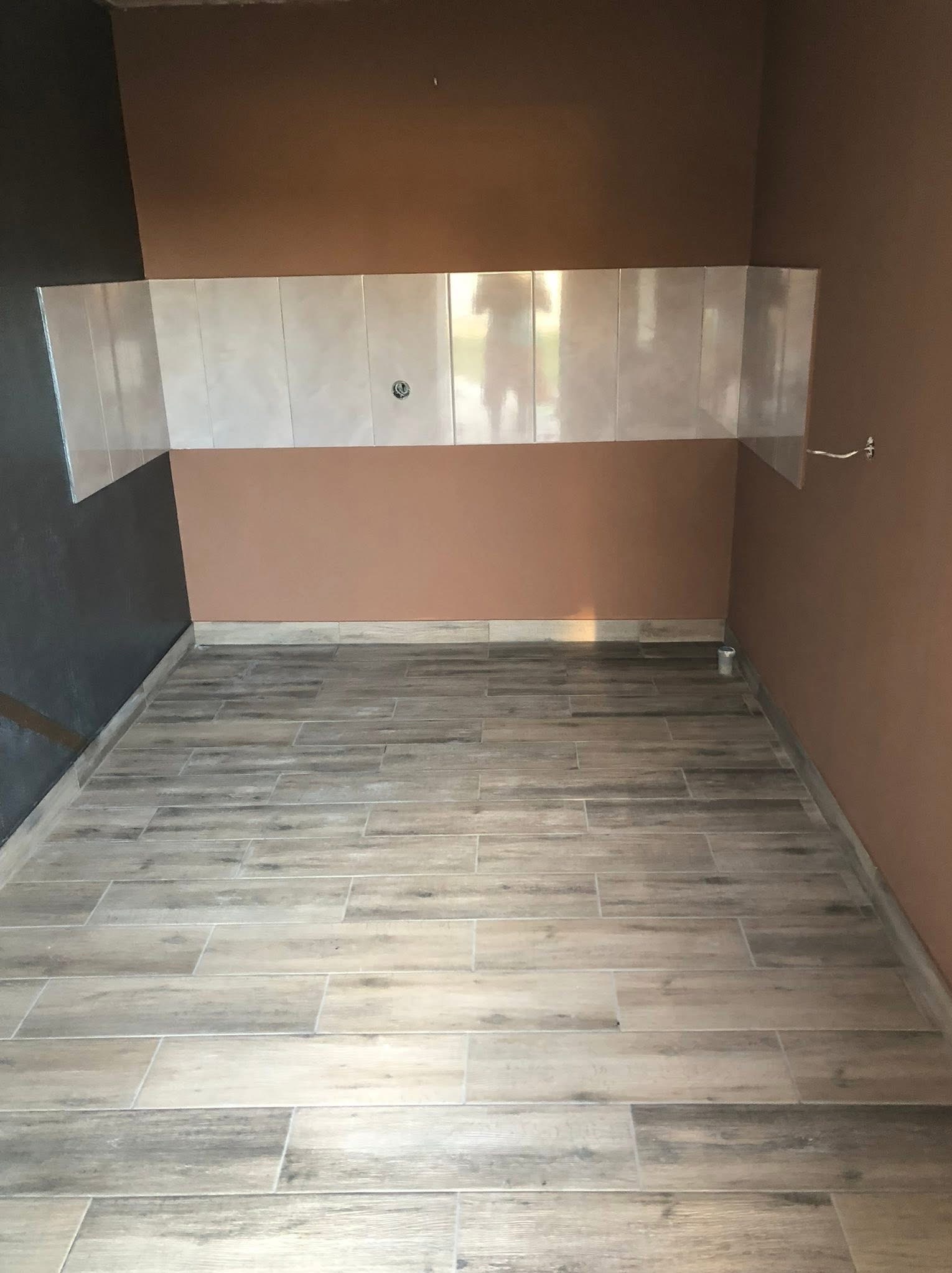 Wykończona łazienka z brązowymi ścianami i ułożonymi płytkami podłogowymi imitującymi drewno. Częściowo ułożone płytki ścienne nad umywalką. Widoczny fragment instalacji.